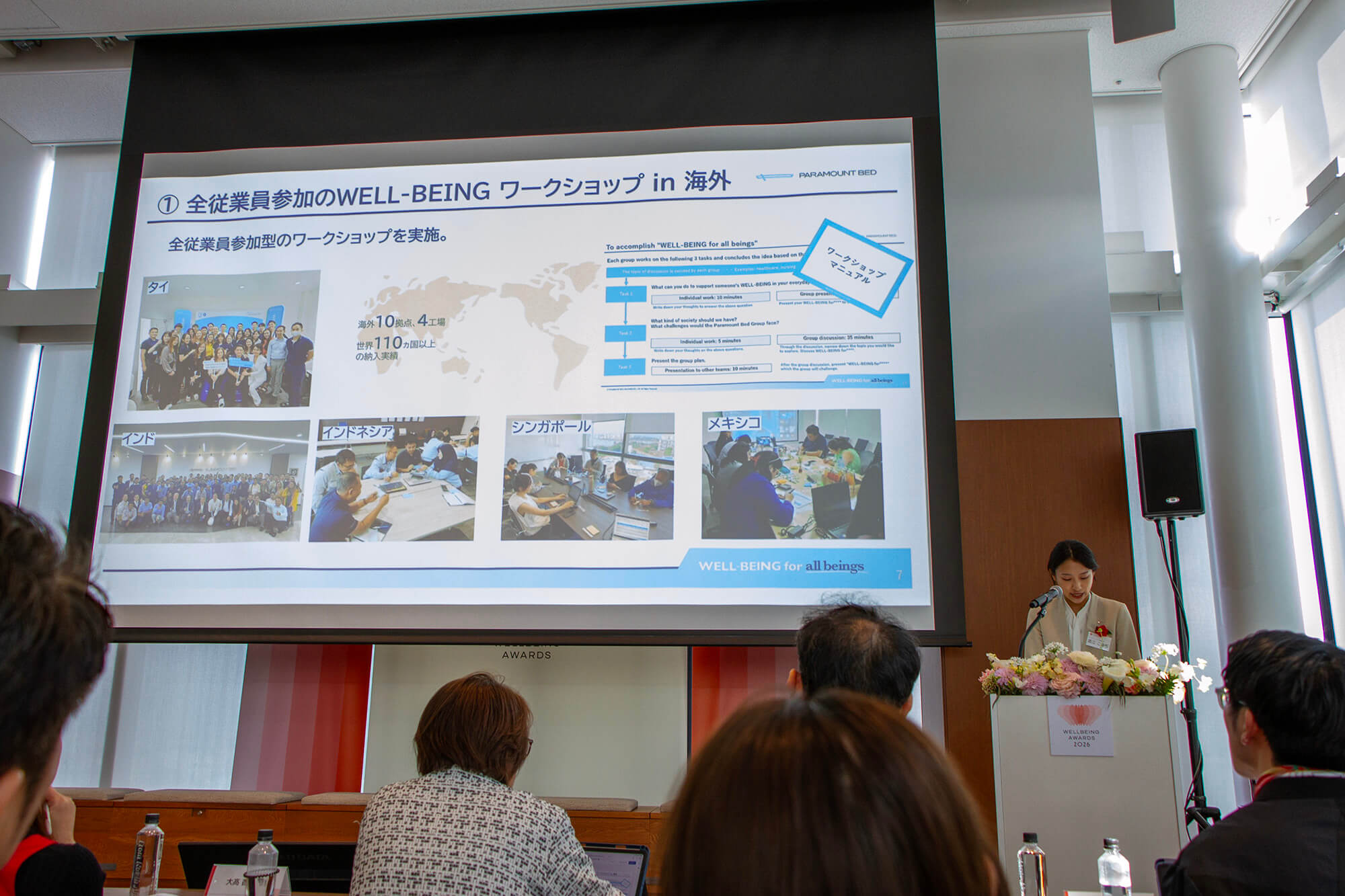 パラマウントベッド株式会社のプレゼンテーション。会場スクリーンには「全従業員参加のWELL-BEINGワークショップ in 海外」の事例紹介